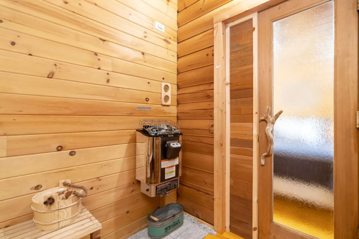 A tranquil, wood-paneled sauna interior featuring a wooden bench, heating unit, bamboo bucket, and frosted glass door. The warm tones and soft lighting evoke a spa-like atmosphere, inviting guests to relax and rejuvenate.