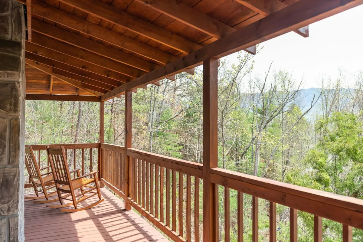 This image features wooden rocking chairs on a deck with a wooden railing, surrounded by trees and framing a picturesque mountain view. The setting exudes peacefulness and invites guests to relax, enjoy morning coffee, or stargaze at night.