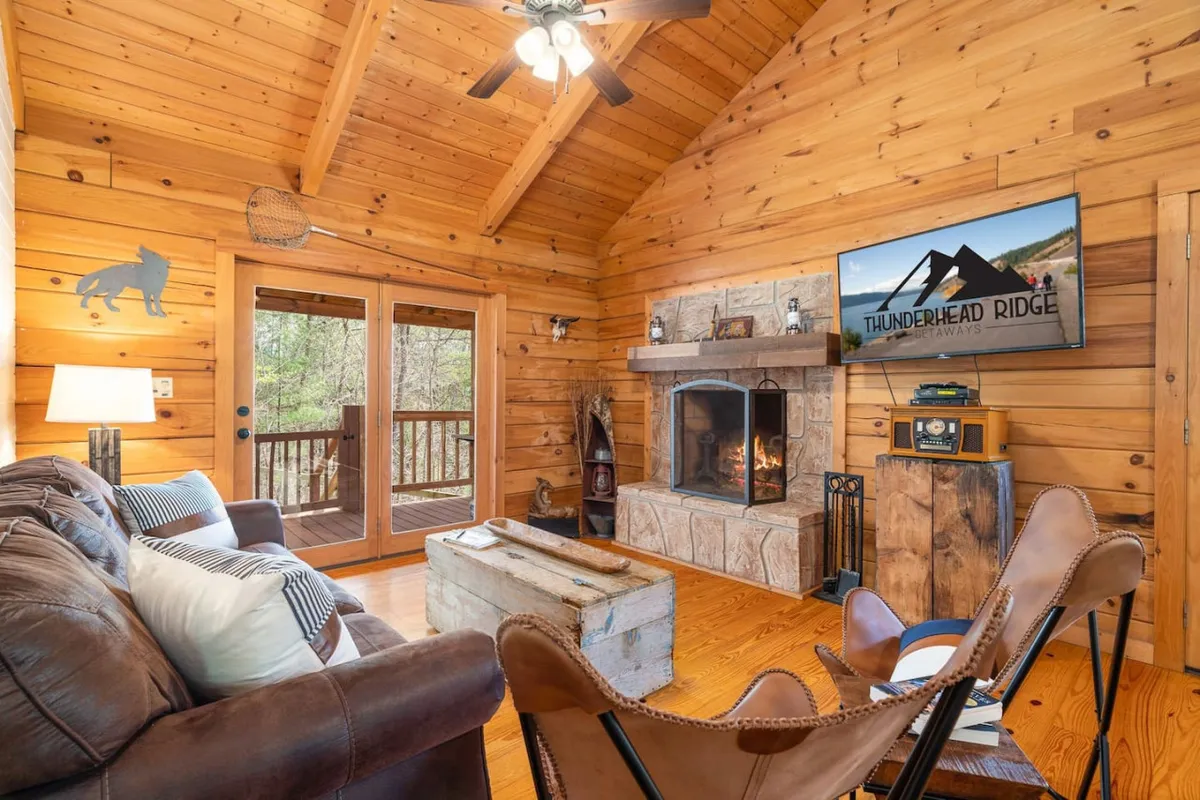 A spacious living area featuring a sofa (which can convert into a queen bed), a beautiful fireplace with wooden beams above, large windows filling the room with natural light, and a TV for entertainment. The room feels warm, inviting, and flexible for different guest needs.