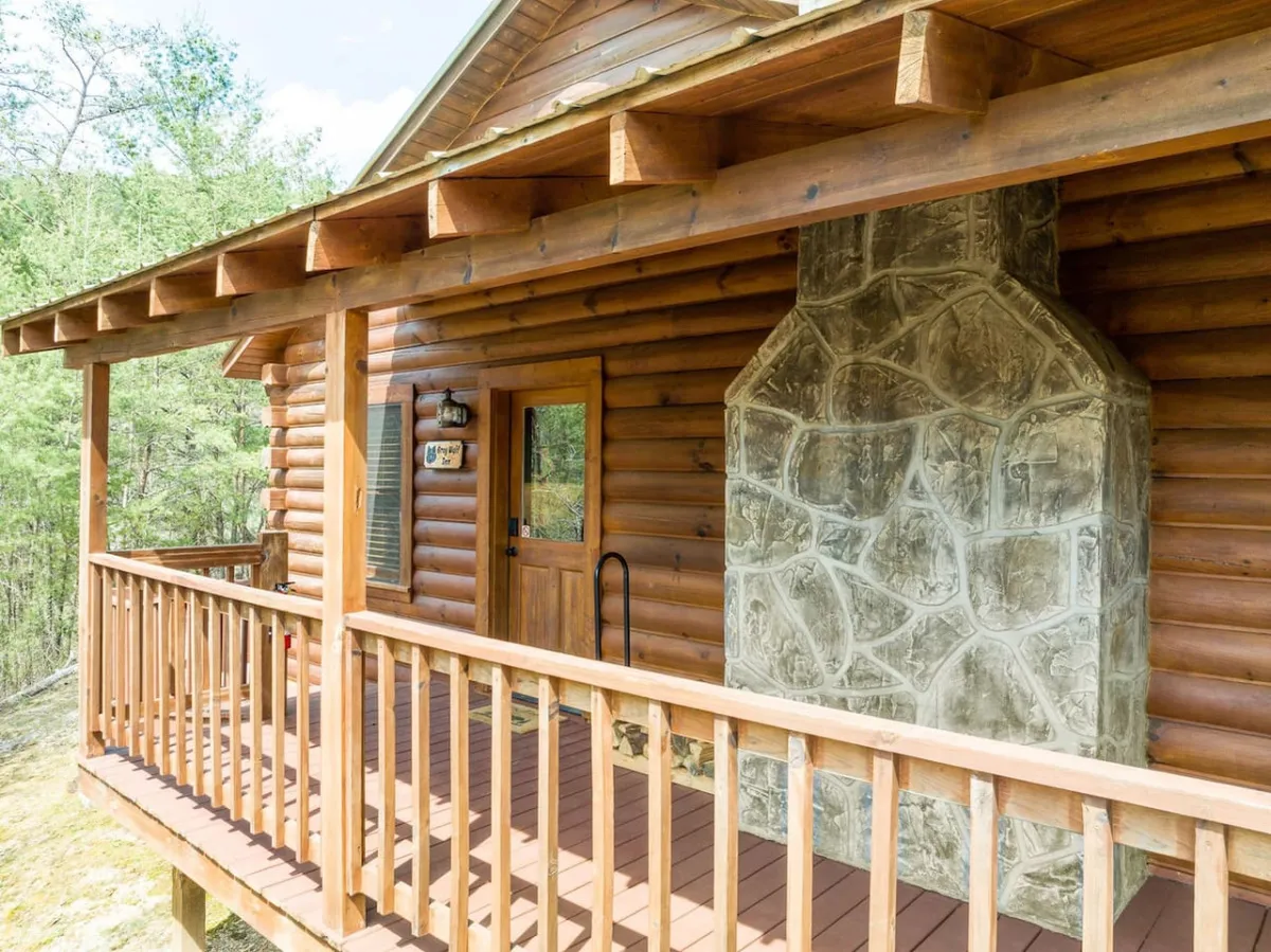 A cozy log cabin exterior with a stone chimney, wooden railing, and welcoming front porch set amidst lush greenery. The inviting look highlights the lodge's rustic charm and connection to the natural surroundings.