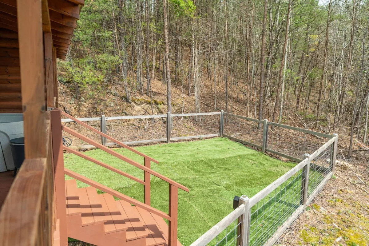 A green artificial grass area bordered by a wooden deck sits amidst a lush forest backdrop. The space is open and inviting, clearly designed for outdoor activities and play. The grassy expanse is perfect for letting pets run and play safely, while the wooden stairs provide easy access between the yard and the deck.