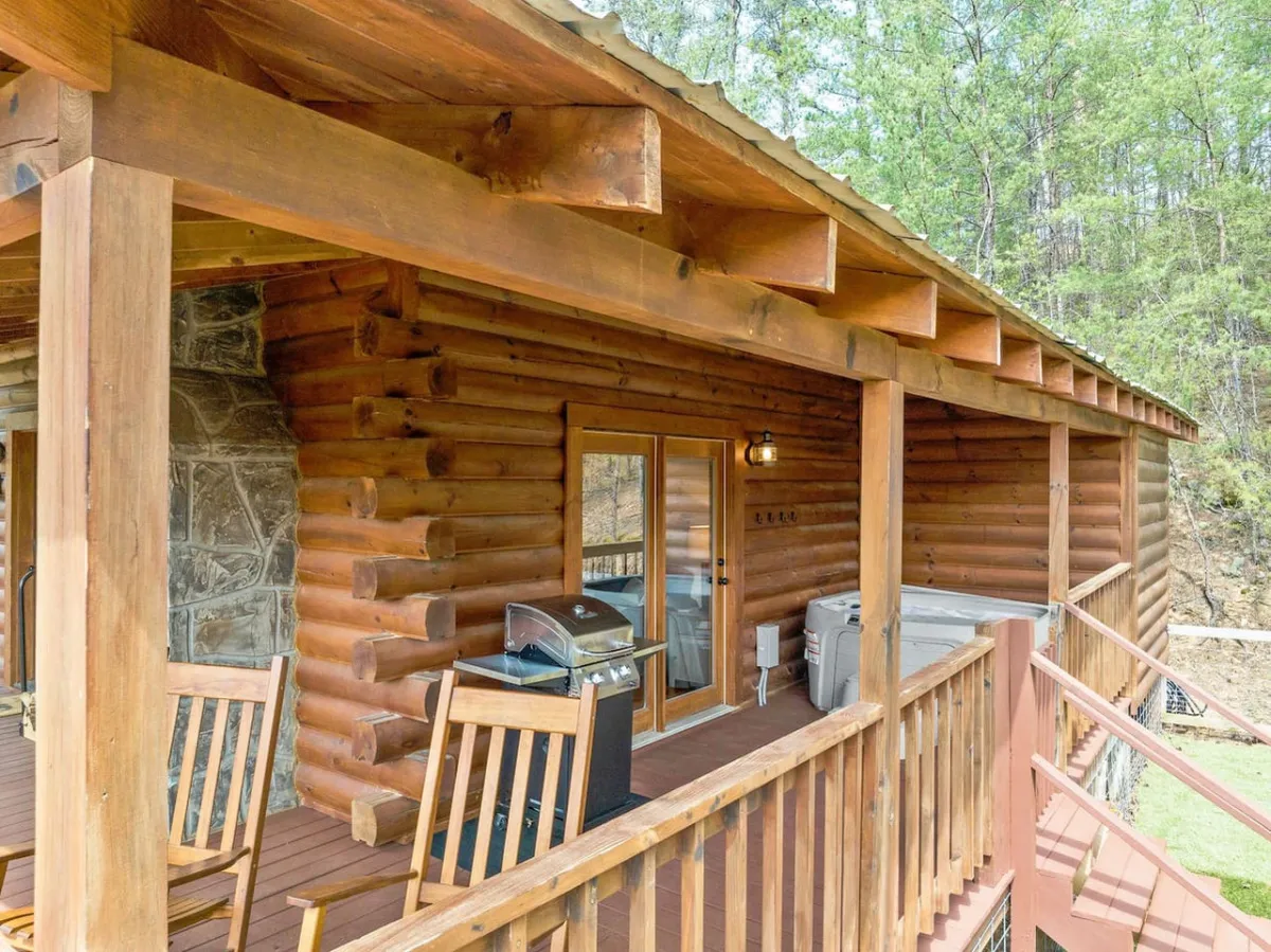 The image shows the log cabin’s exterior, featuring a welcoming deck with outdoor seating and a grill. Rock accents and abundant greenery surround the home, creating an atmosphere ideal for outdoor meals, morning coffee, or evening relaxation.