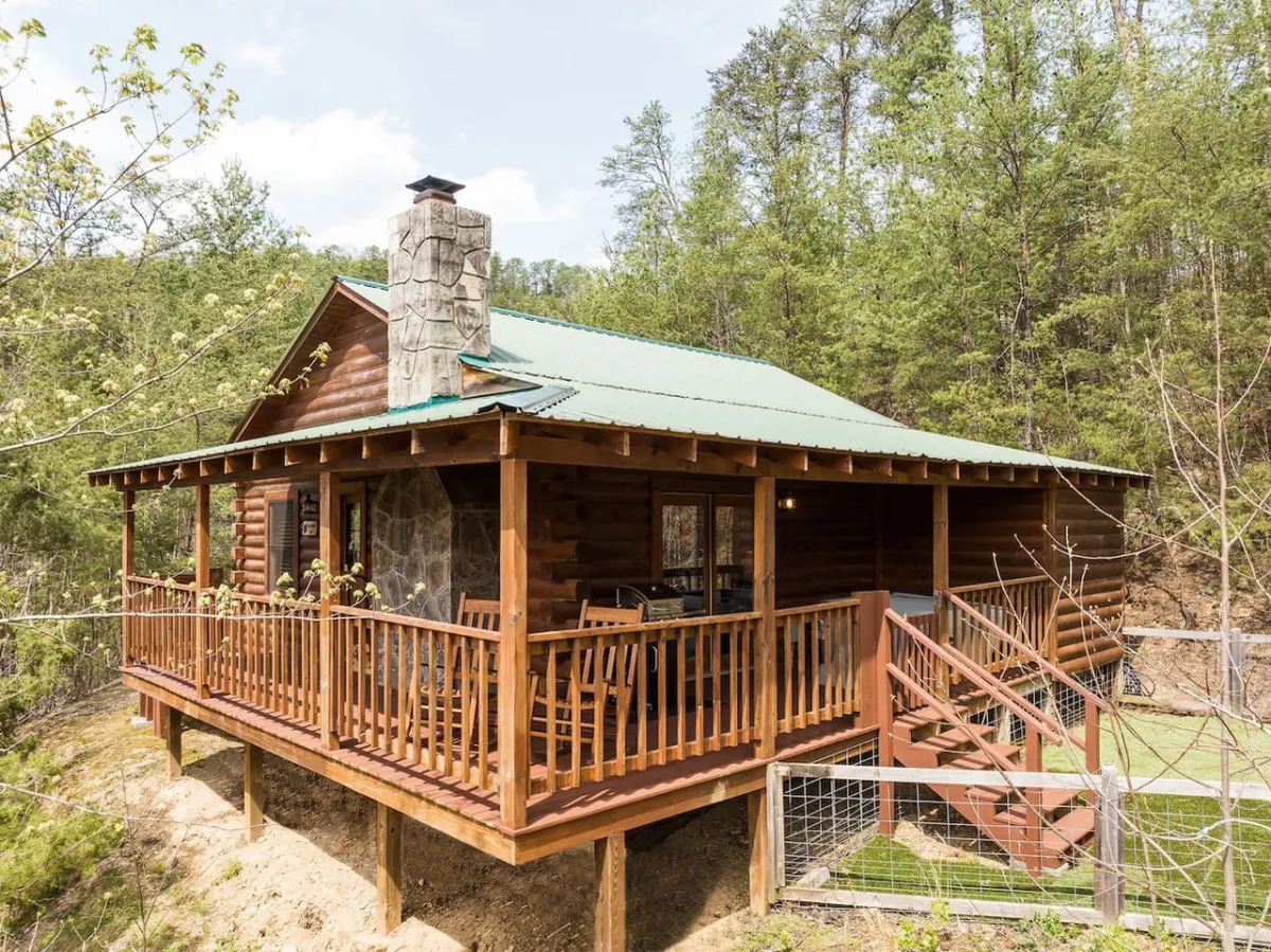 The image showcases the warmly lit exterior of a renovated wooden cabin nestled among lush trees, featuring a striking stone chimney, a picturesque green roof, and a spacious porch perfect for relaxation and gathering. The setting radiates coziness and tranquility, with inviting outdoor seating where guests can savor mountain sunsets, morning coffee, or peaceful stargazing.