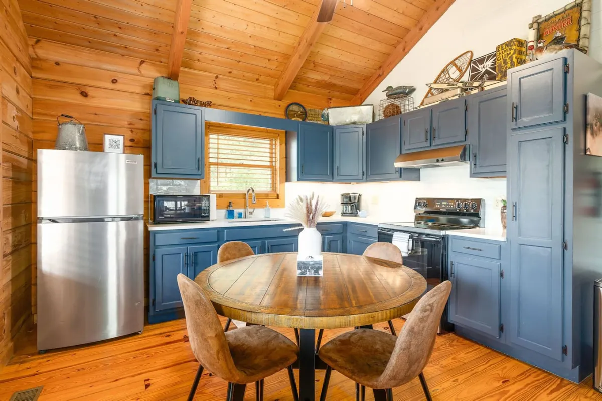 This scene spotlights the round wooden dining table surrounded by comfortable chairs, set against blue cabinetry and stainless steel kitchen appliances. The space feels cozy and ideal for shared meals or celebrations, with ample natural light.