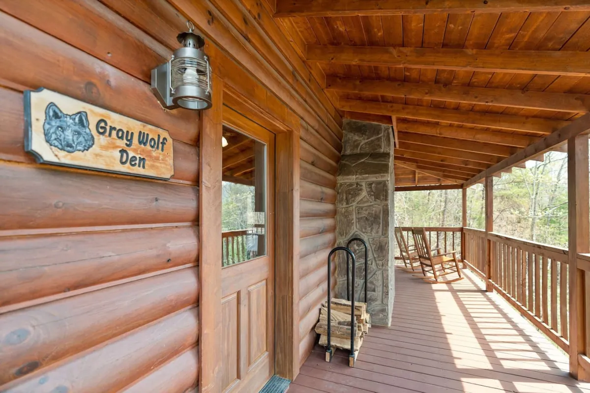 This image shows the entrance to Gray Wolf Den, featuring a welcoming wooden door, rustic stone wall, and inviting rocking chairs on the porch. The prominent 'Gray Wolf Den' sign clearly marks the cabin, and the surrounding natural scenery hints at an outdoor-friendly setting.