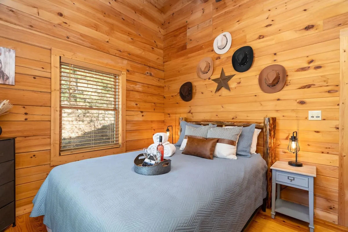 A comfortable, rustic bedroom with wooden wall panels, a bed adorned with decorative pillows, hats displayed as wall art, and a nightstand with a cozy lamp. The ambiance is restful and homely, highlighting the peaceful charm of the bedroom.