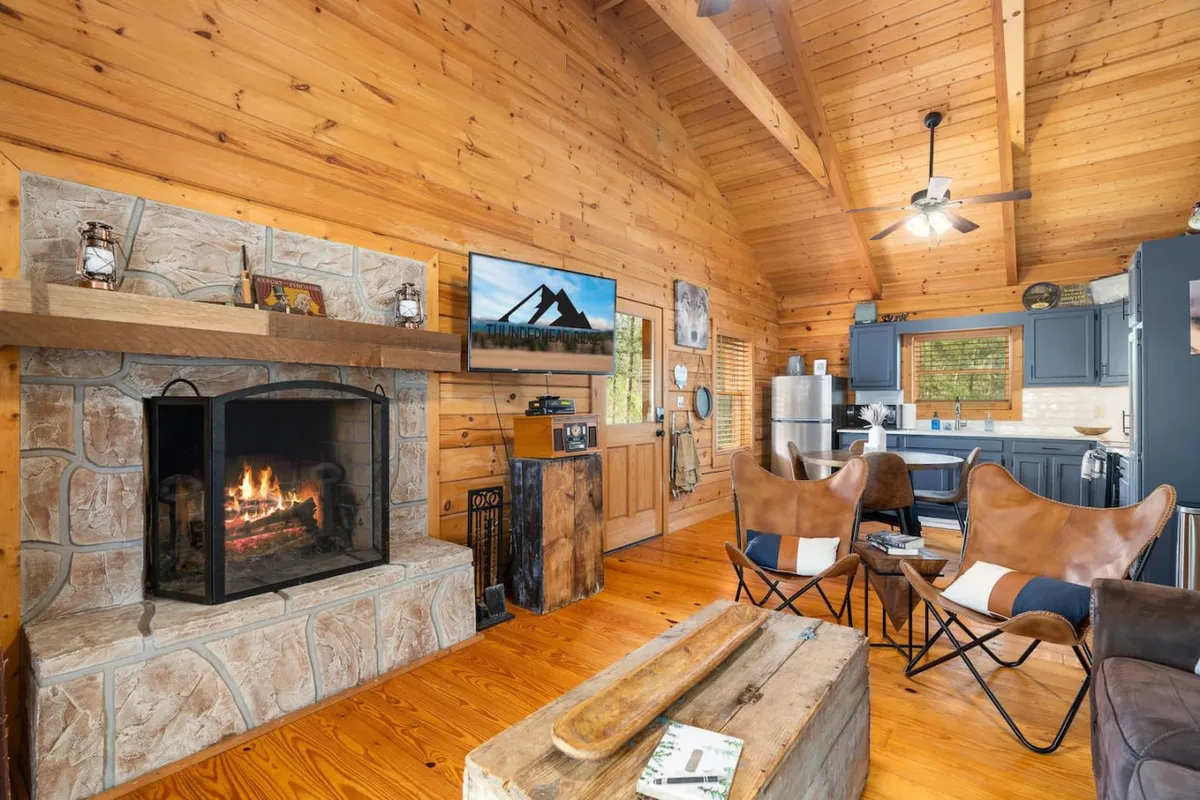 This image captures the open-concept living area, featuring a real-wood fireplace, exposed wooden beams, plush seating, a TV, and a glimpse of the adjacent kitchen. The cozy textures and warm lighting evoke a welcoming mountain retreat atmosphere.