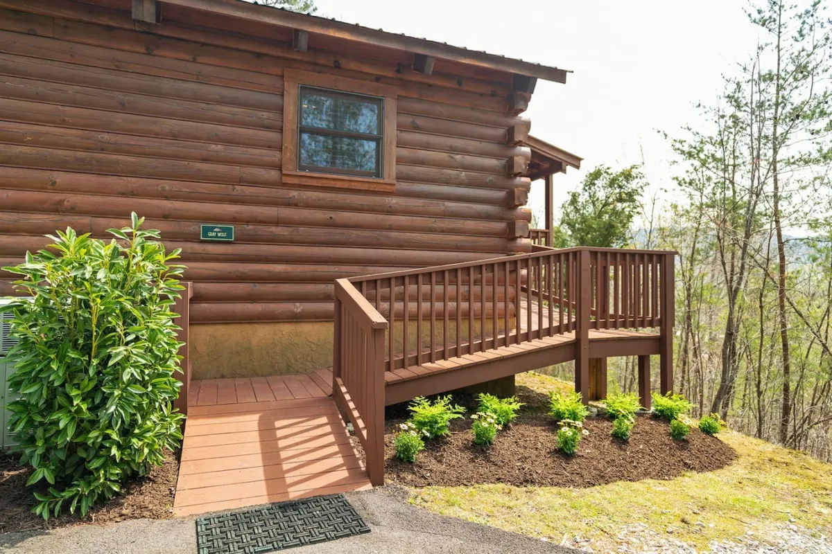 This image features the log cabin structure of Gray Wolf, set within lush greenery, and prominently displays a wooden ramp leading to a step-free entrance. The inviting porch is ideal for relaxing or gathering with family, while the surrounding trees emphasize the privacy and natural seclusion of the gated community.