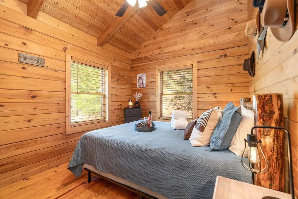 A cozy bedroom featuring a plush bed with a blue quilt, warm wooden walls, windows with blinds letting in soft natural light, and decorative hats adding a personal touch. The space radiates tranquility and comfort, providing a restful atmosphere for relaxation.